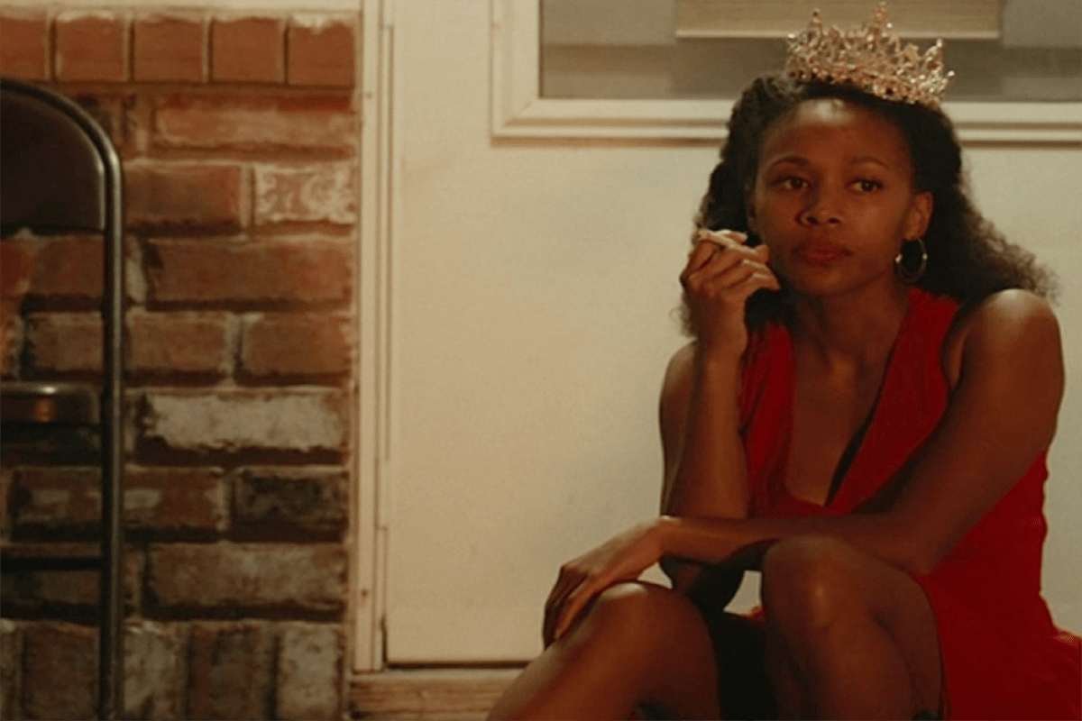 A woman sits smoking, wearing a tiara and red dress, against a brick wall and door.