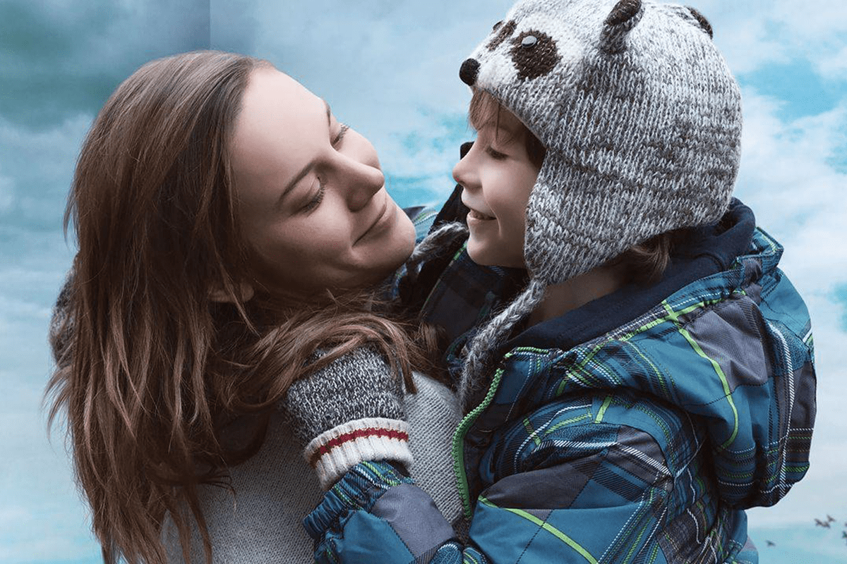 Woman joyfully holds smiling child in a bear hat, against a cloudy sky background.