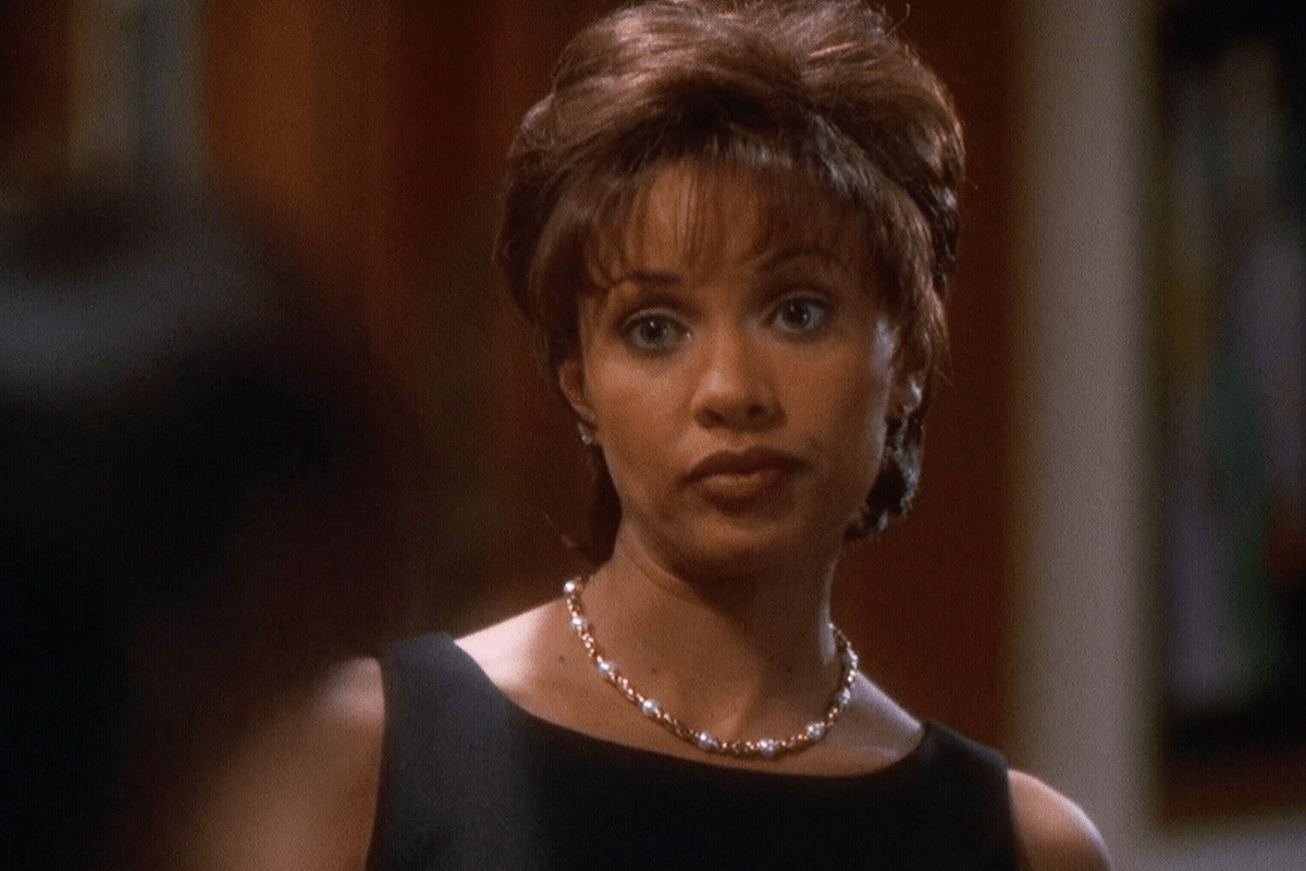 A person wearing a pearl necklace looks forward, standing indoors against a softly blurred background.