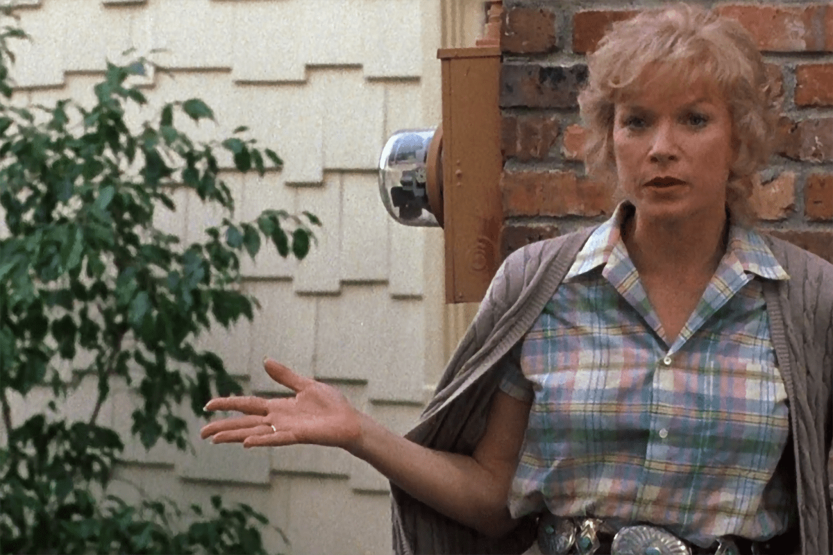 Woman gestures with hand, standing against a brick wall near foliage, wearing a plaid shirt and cardigan.