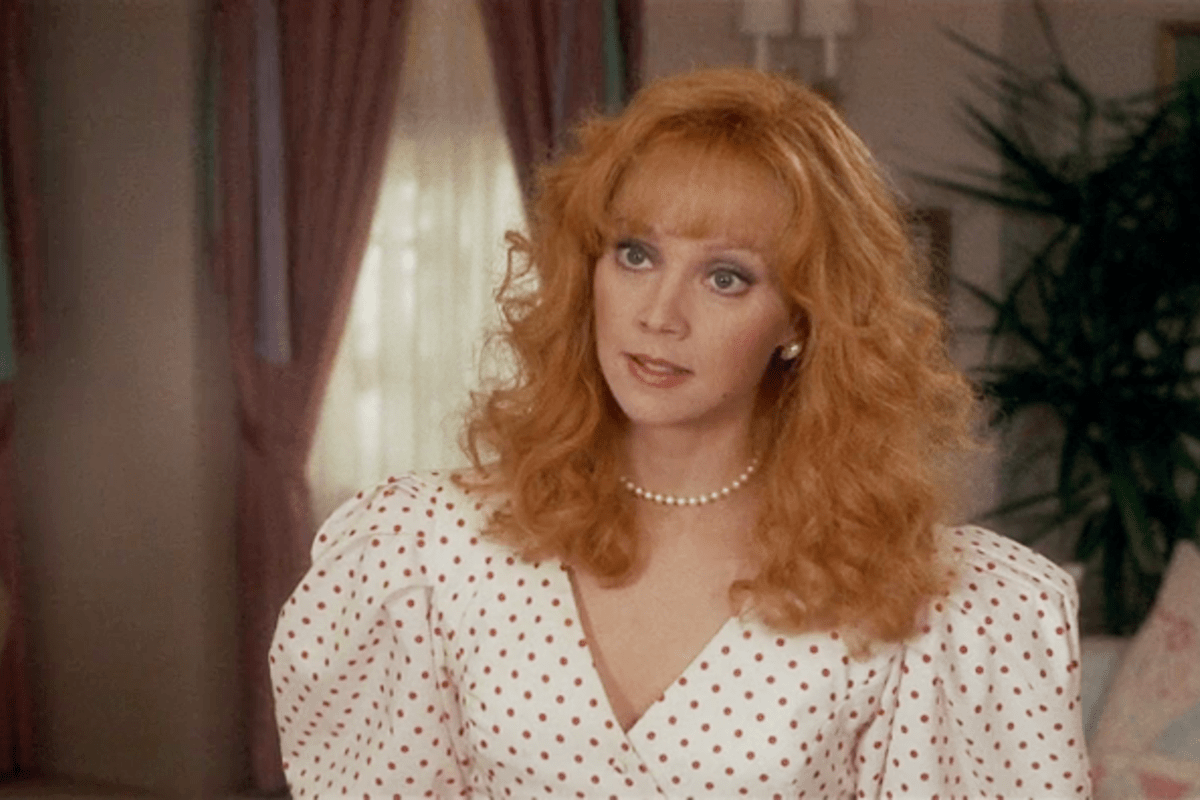 Woman with curly red hair and pearl necklace stands indoors wearing white dress with red polka dots; curtains and plant in the background.