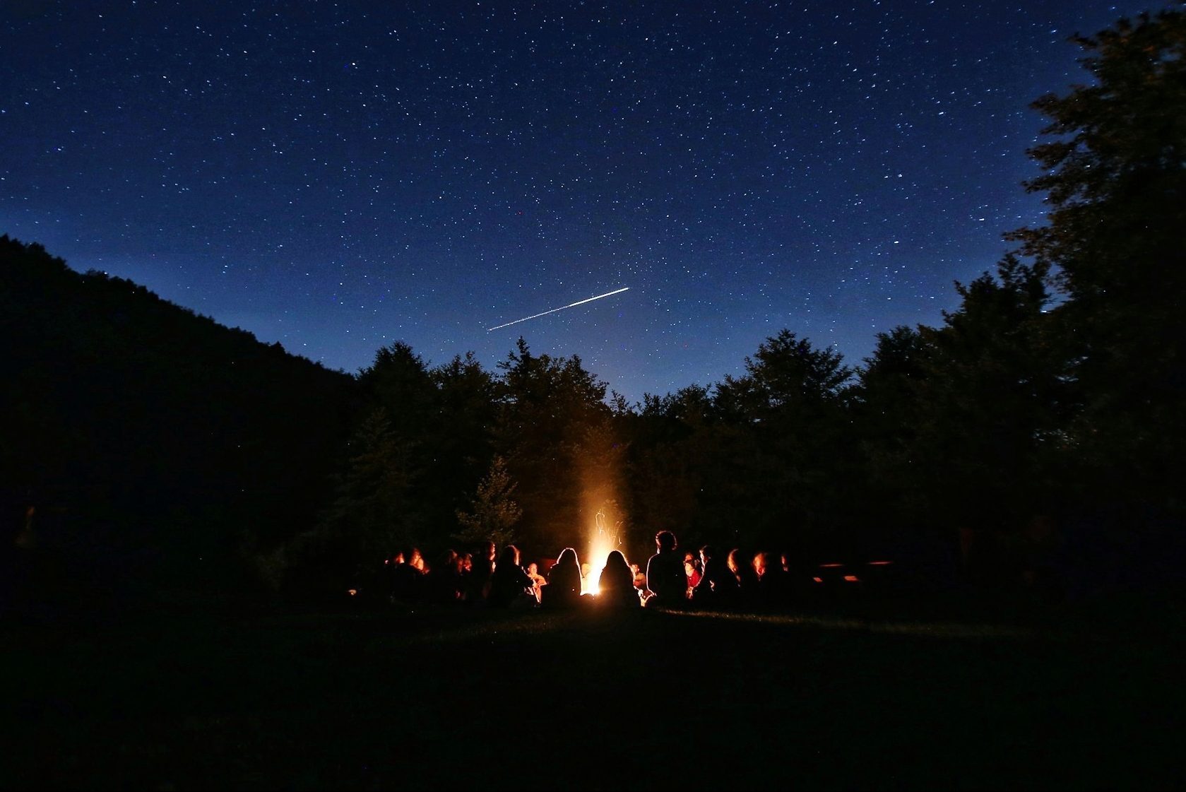 People sit around a campfire beneath a starry sky, with a shooting star above and trees surrounding the scene.