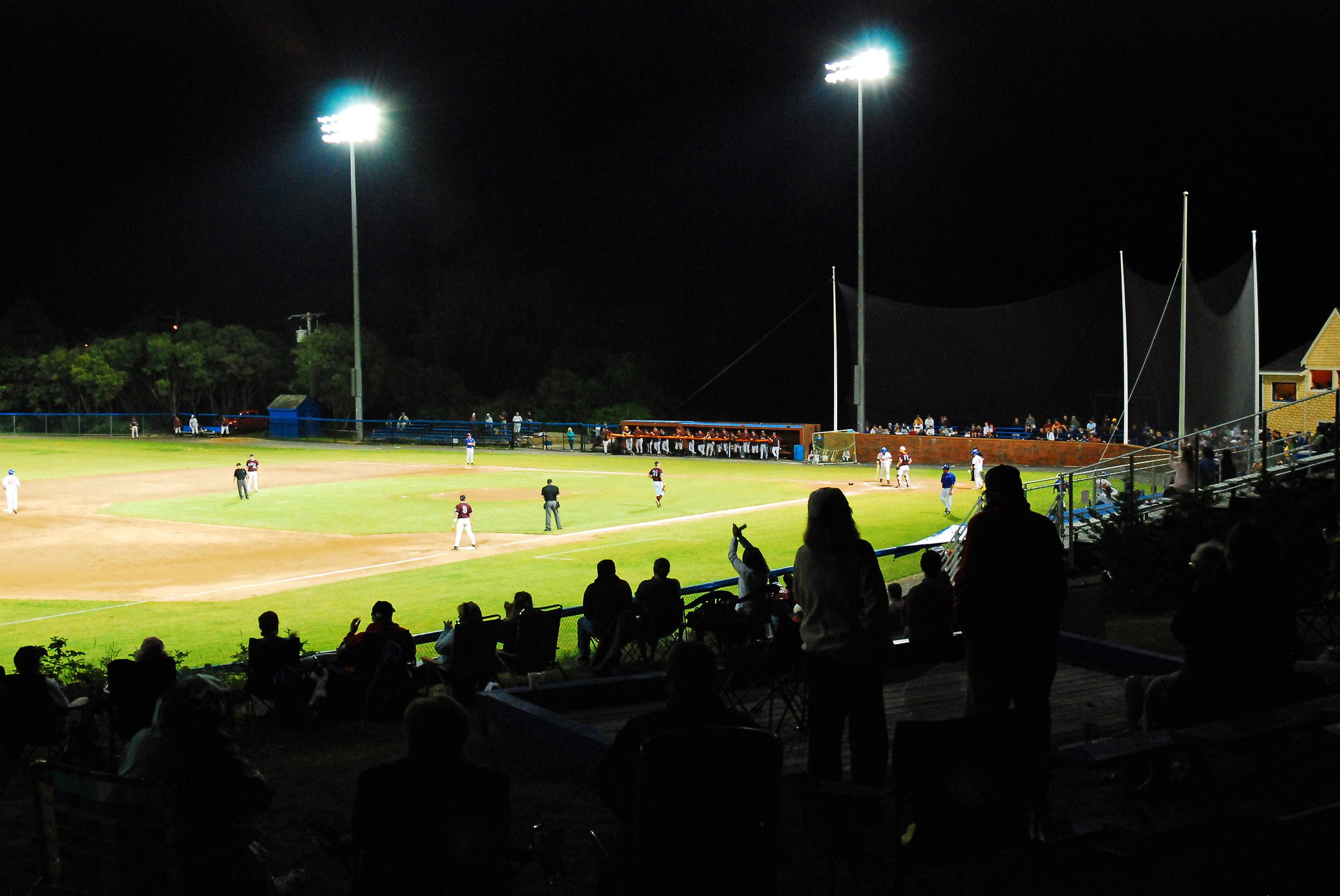 Baseball players compete under bright stadium lights at night, with spectators in silhouette watching from the stands.