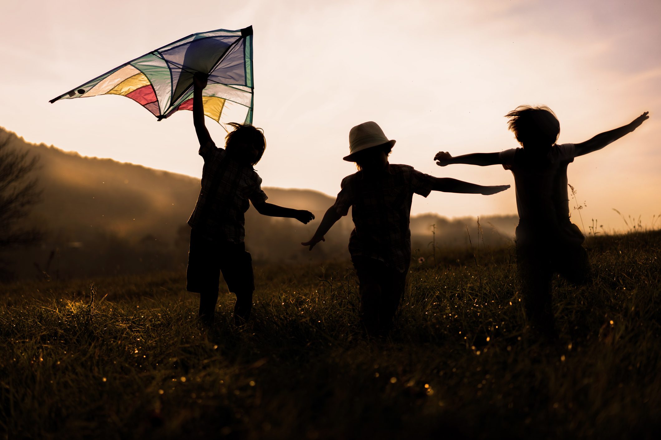 Children run joyfully with a kite in hand, silhouetted against a sunset on a grassy hill.