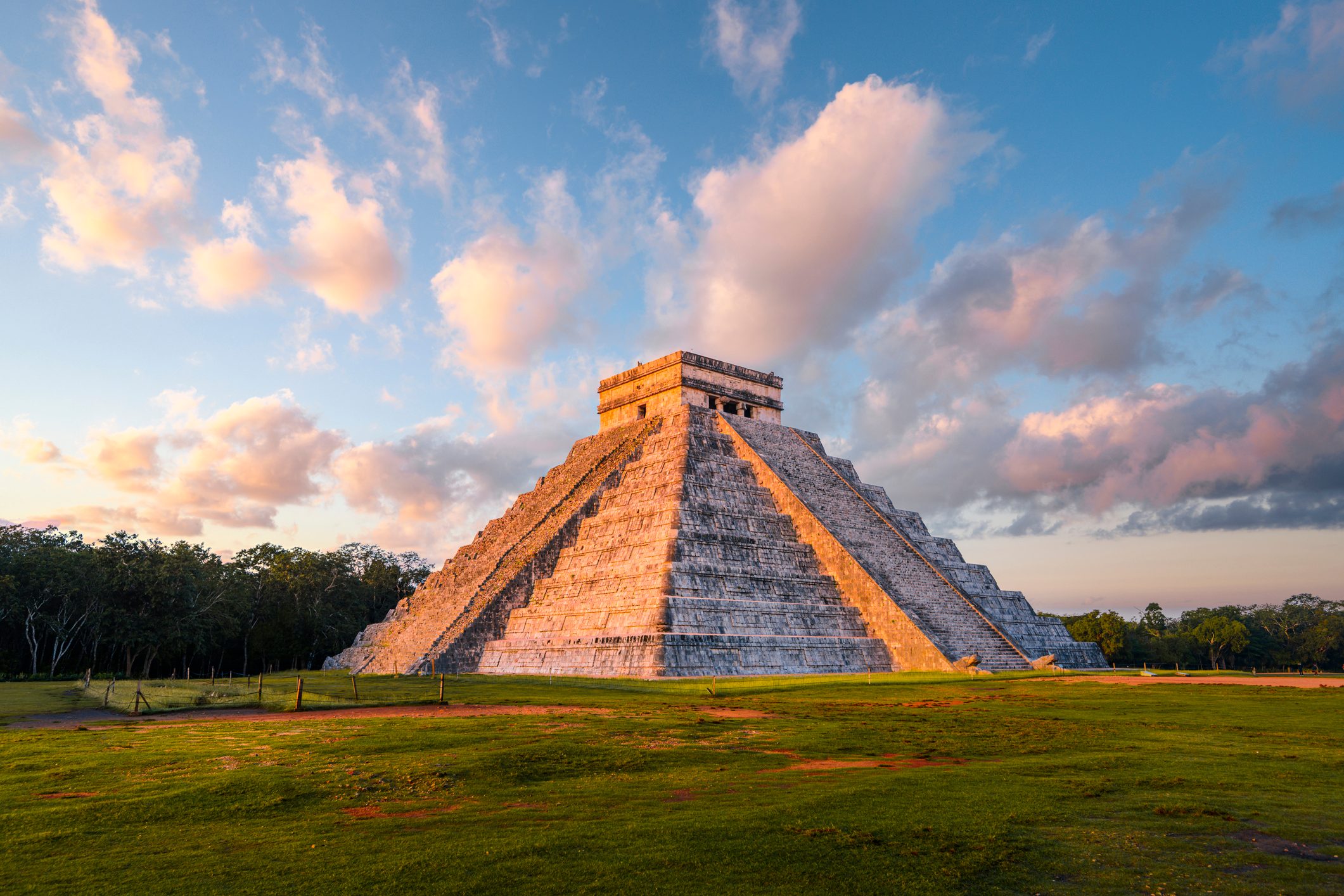 A stepped pyramid stands under a colorful sky, surrounded by green grass and scattered clouds, illuminated by soft, golden sunlight.