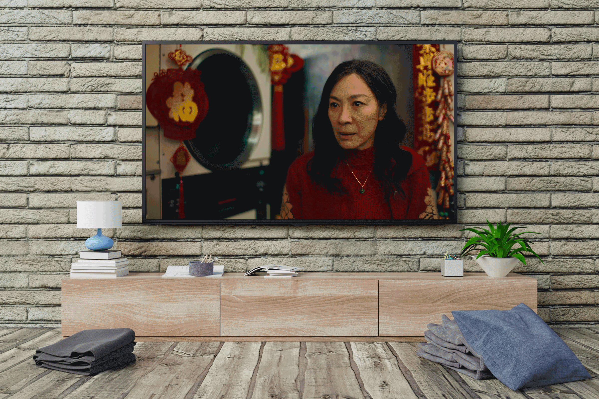 Television shows a focused woman indoors; laundry is visible. The screen is mounted above a wooden console with a lamp, books, plants.