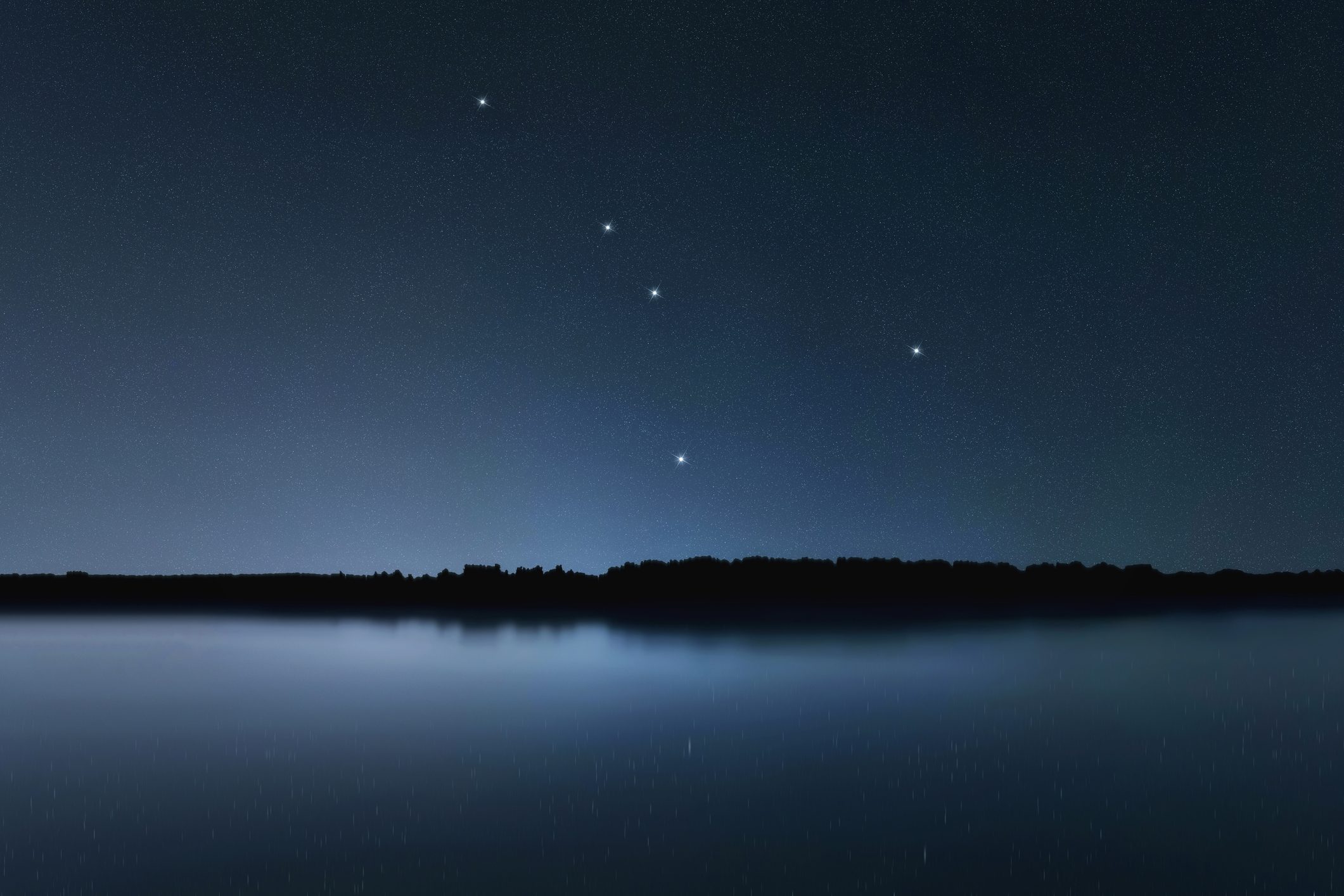 Stars shine brightly in the night sky over a calm lake, reflecting a dark silhouette of distant trees on the horizon.
