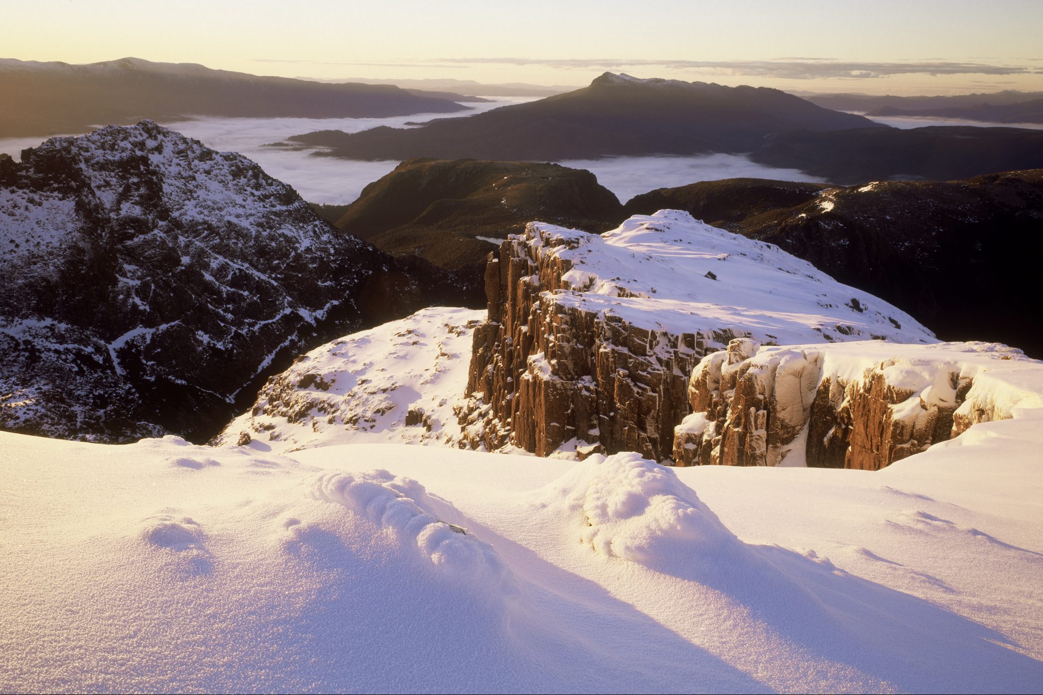 Snow-covered mountain peak illuminated by sunrise, with a rocky cliff and layered mountain ranges under a misty sky in the background.