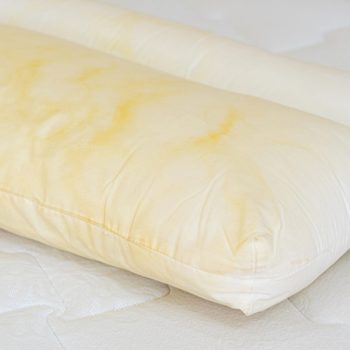 Yellowed pillow lies on a mattress, displaying noticeable stains, within a bedroom setting.