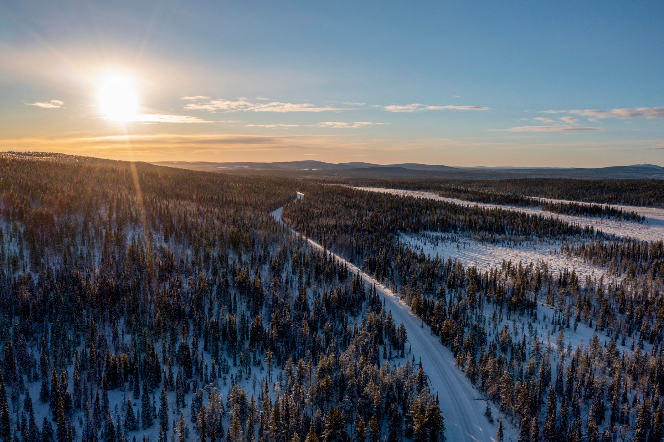 The sun rises over a snow-covered forest, illuminating a winding road through the trees in a vast, wintry landscape.