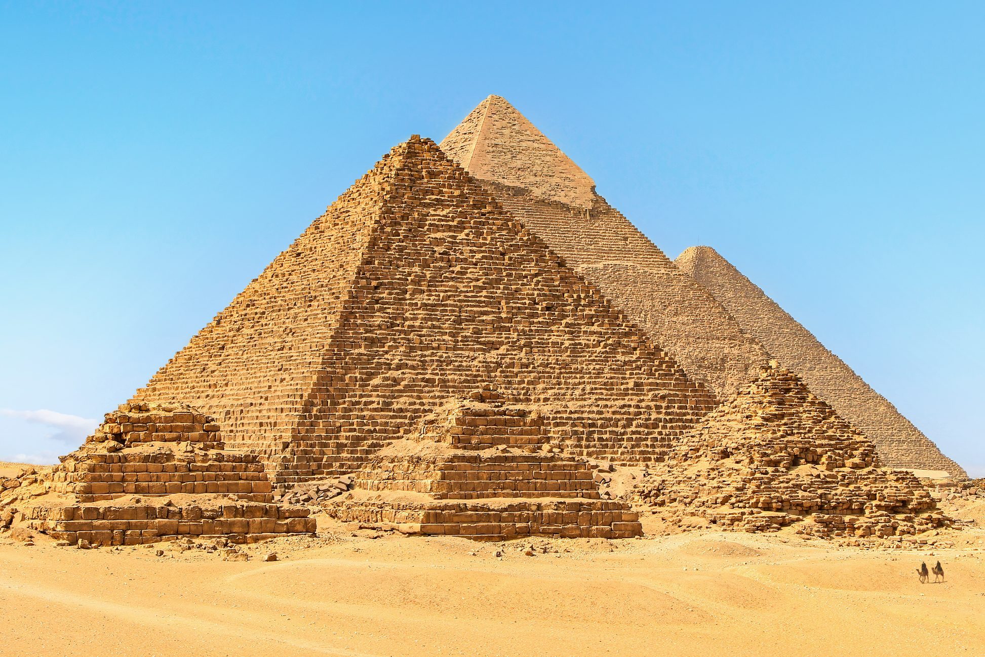 Pyramids stand majestically amidst vast desert landscape under clear blue sky, with two distant camels in the foreground.