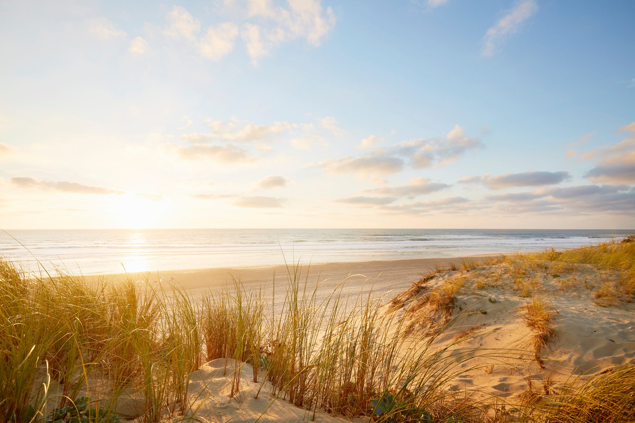 Sun sets over ocean, illuminating sandy dunes with grasses, casting long shadows in the serene beach landscape under a partly cloudy sky.
