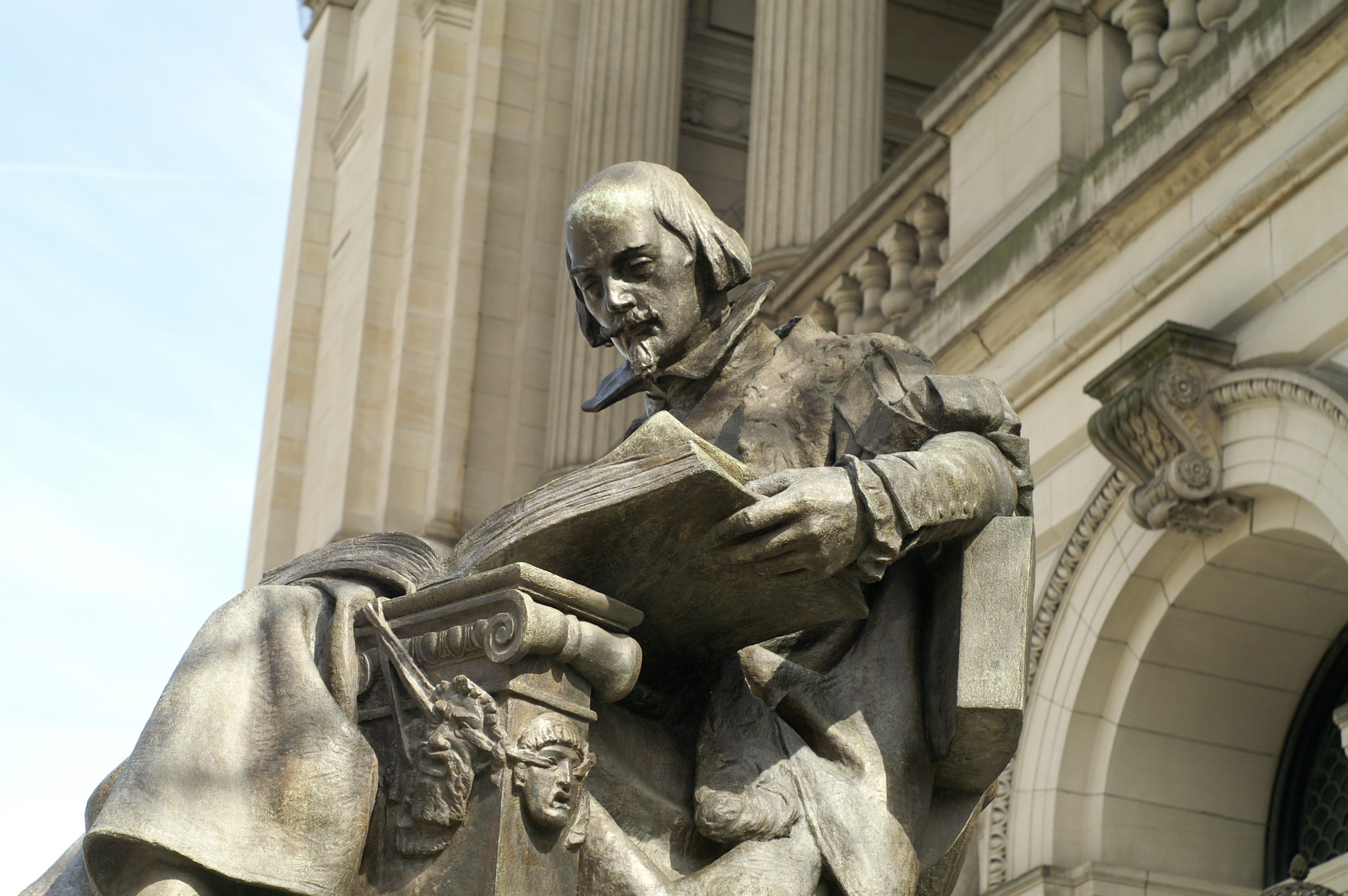 A bronze statue leans over a large book, set against the backdrop of a classical stone building with columns.