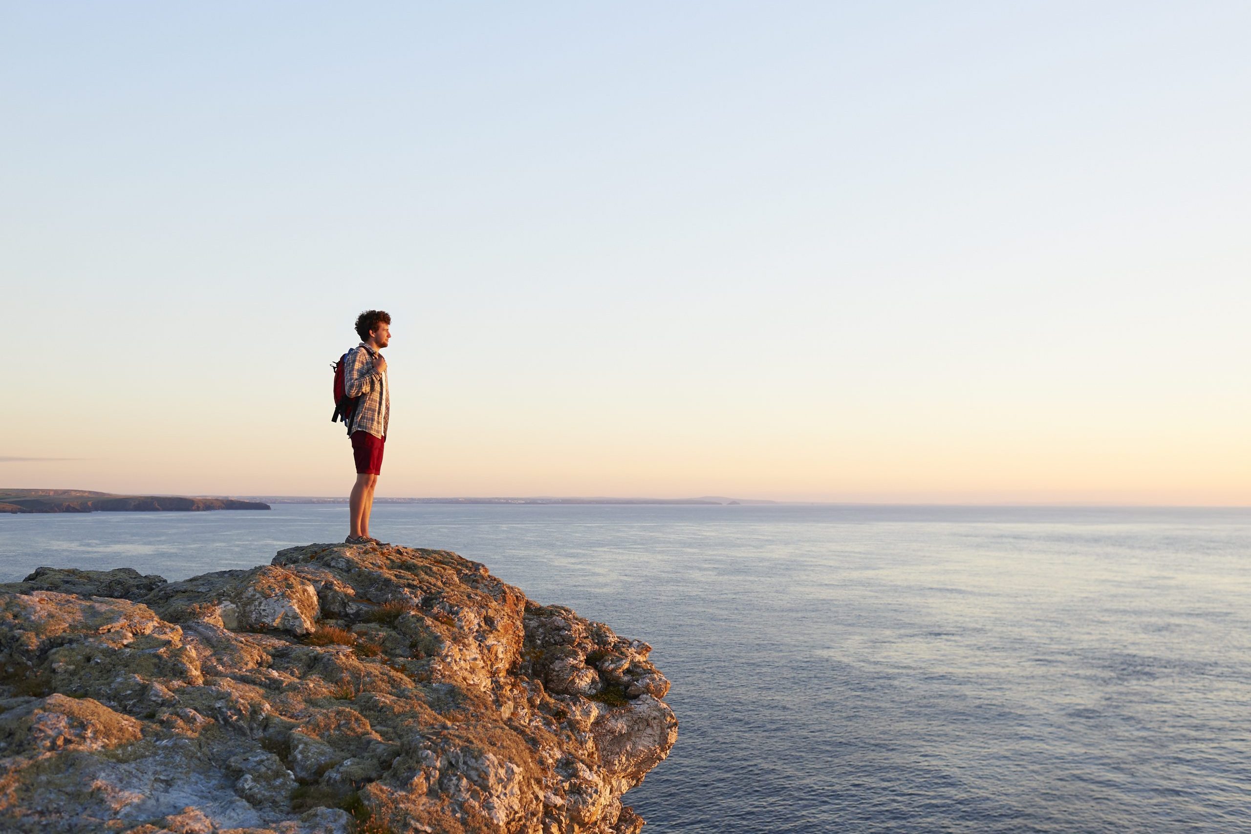 A person stands with a backpack, overlooking the ocean from a cliff at sunset, surrounded by a tranquil sky and distant horizon.