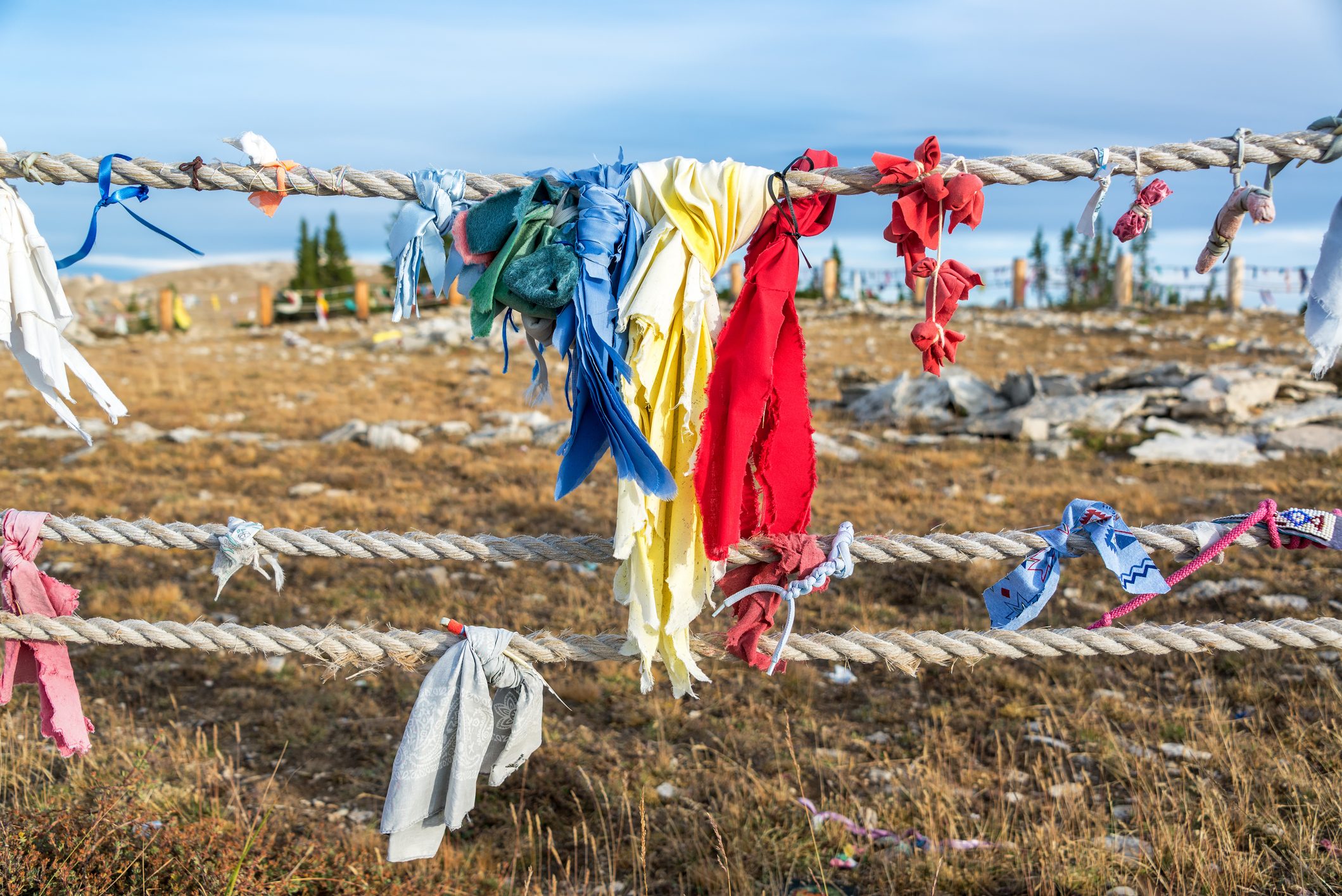 Colorful cloth strips hang from a rope fence in a rocky, grassy landscape under a clear sky.