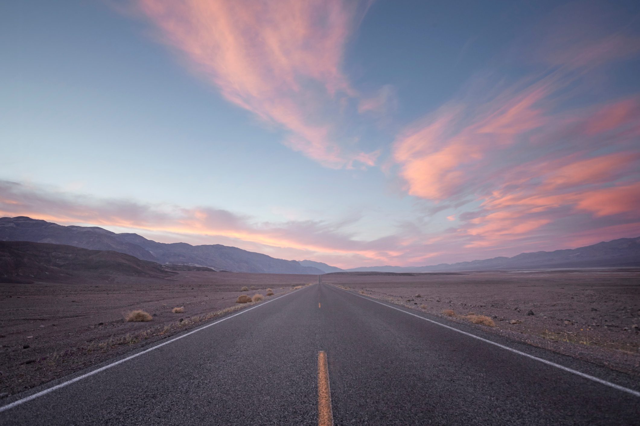 Road stretches into desert landscape, framed by pink-tinged clouds in a vast, open sky.