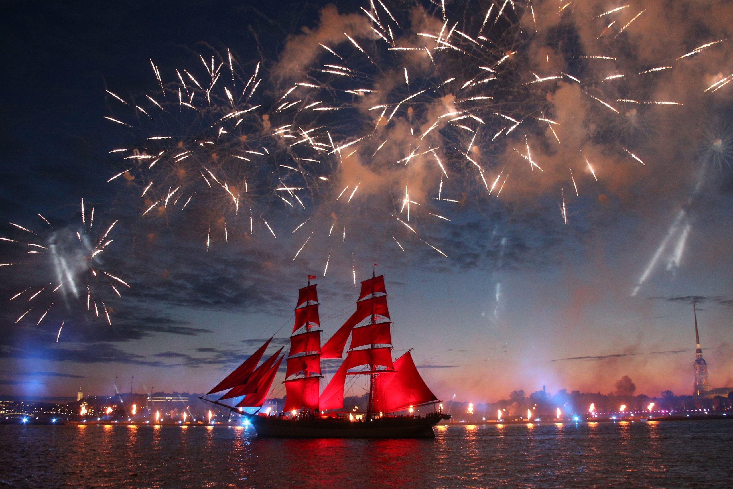 Sailing ship moves beneath vibrant fireworks, illuminating the night sky over a cityscape by the water.