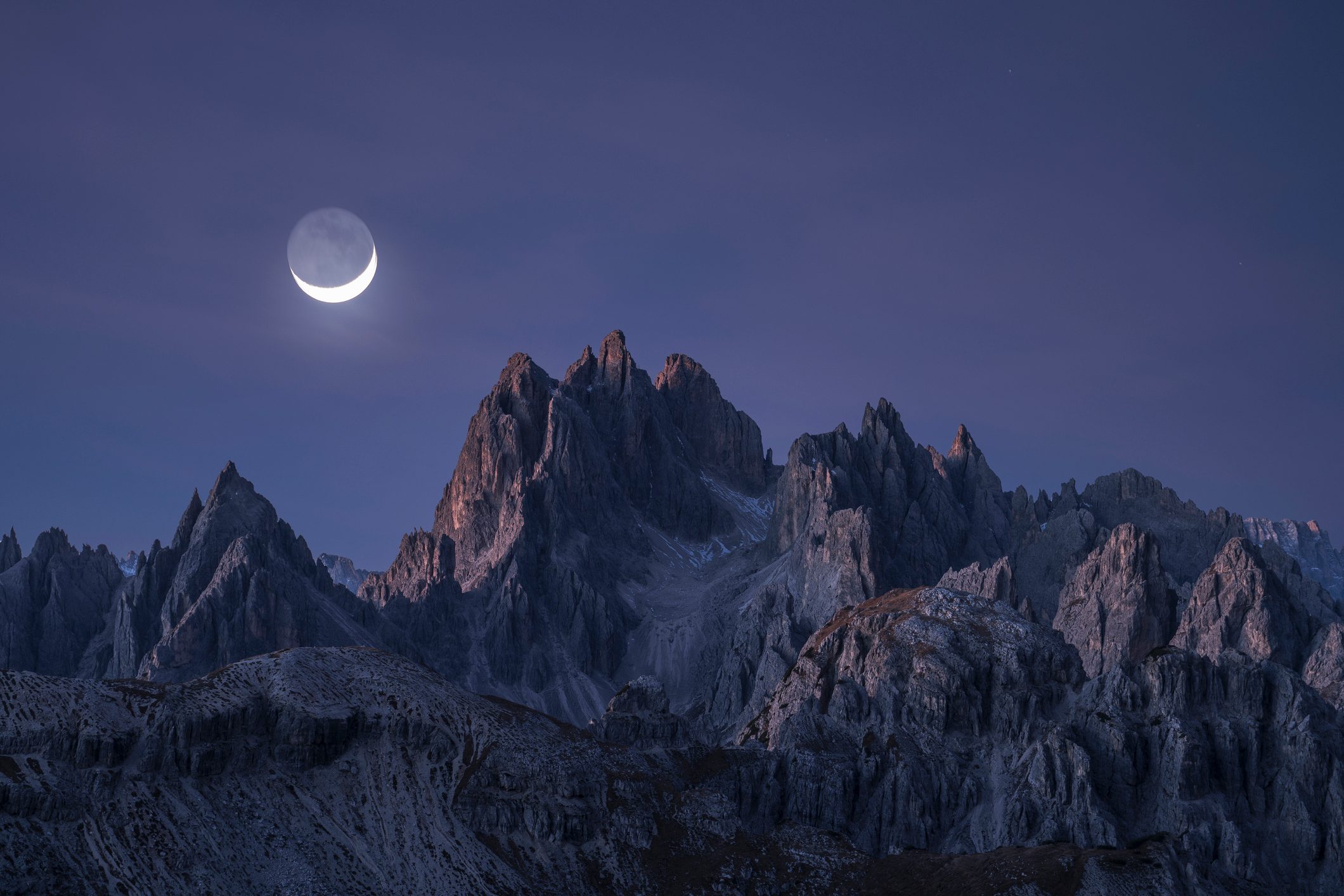 Crescent moon shines over jagged mountain peaks during dusk under a clear, darkening sky.