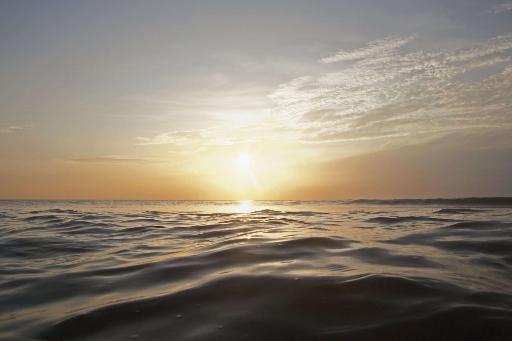 Sun sets over rippling ocean waves, creating a warm, golden sky with scattered clouds above the horizon.