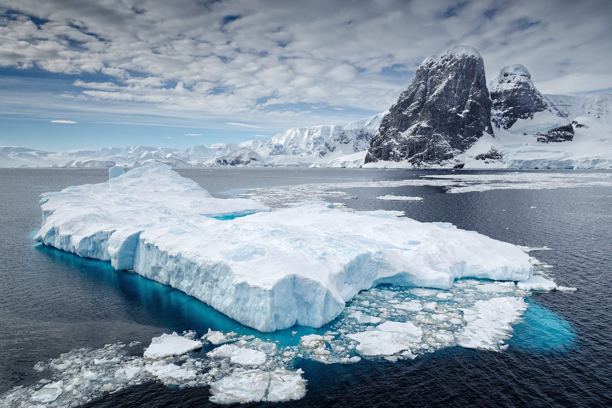 Iceberg floats serenely; snowy peaks and calm, expansive ocean surround under a cloudy blue sky.