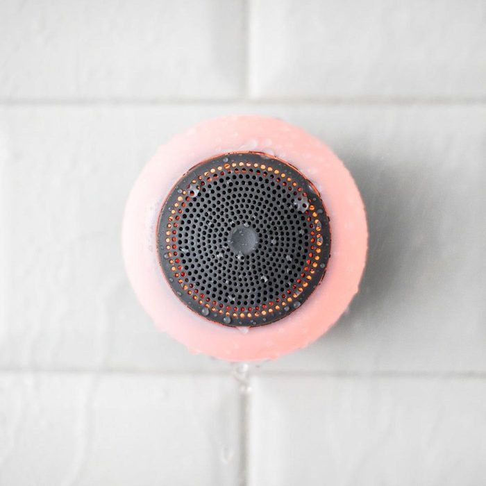 Round speaker faces viewer, placed against wet, tiled surface, with water droplets; pink casing and perforated black front with orange detail.