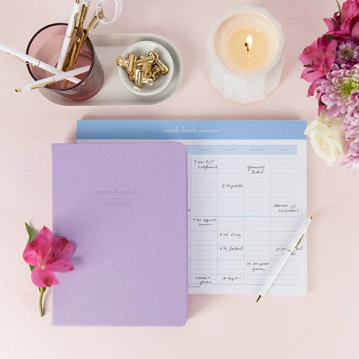 Planner sits open with handwritten notes; surrounded by a pink notebook, lit candle, pens in a glass, pink flower, and gold clips.
