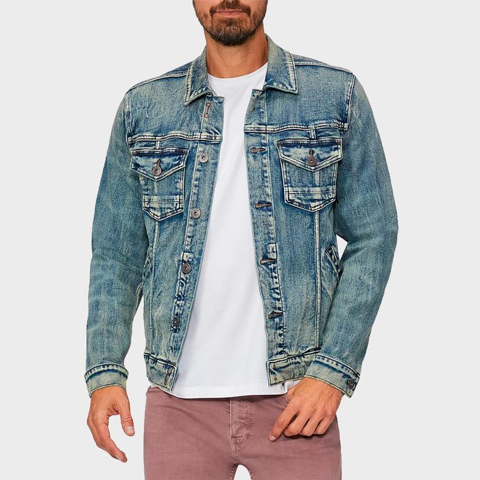 A man wears a blue denim jacket over a white t-shirt and pink pants, standing against a plain background.