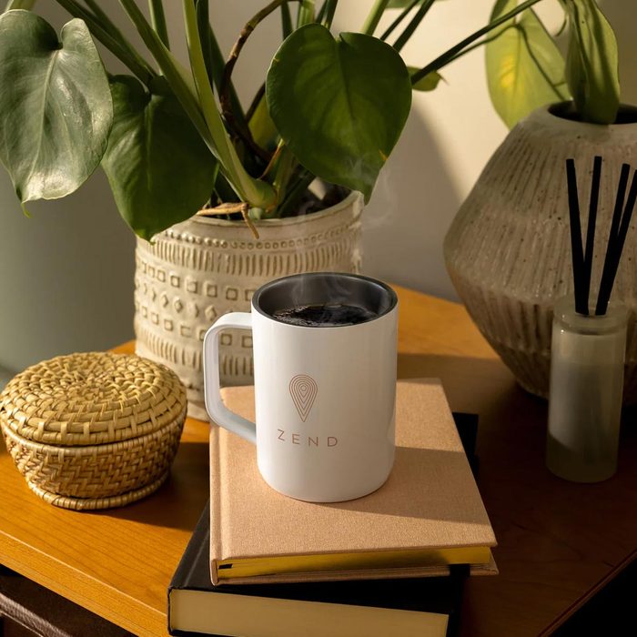 White mug emitting steam sits on a book in a cozy setting with plants and decor. 
