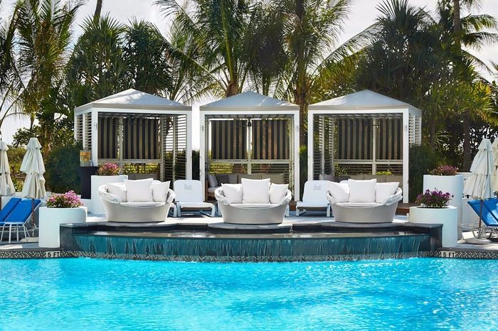 Poolside seating includes round chairs and cabanas, surrounded by tropical palm trees and loungers, near a sparkling blue swimming pool with cascading water.