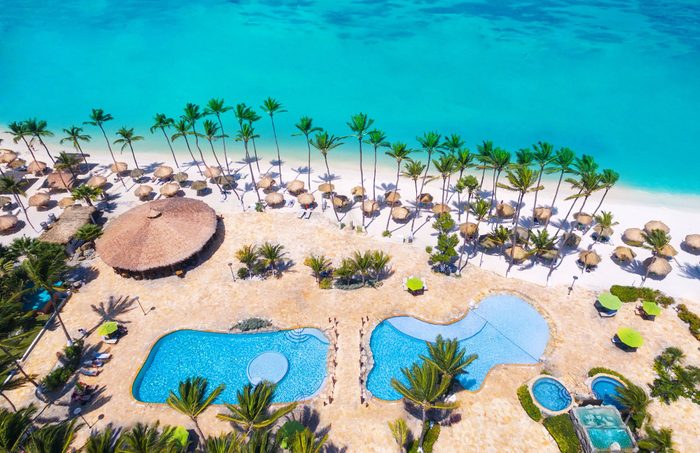 Pools shimmer by palm tree-lined beachfront, surrounded by sun loungers and thatched umbrellas, adjacent to a circular, open-air pavilion.