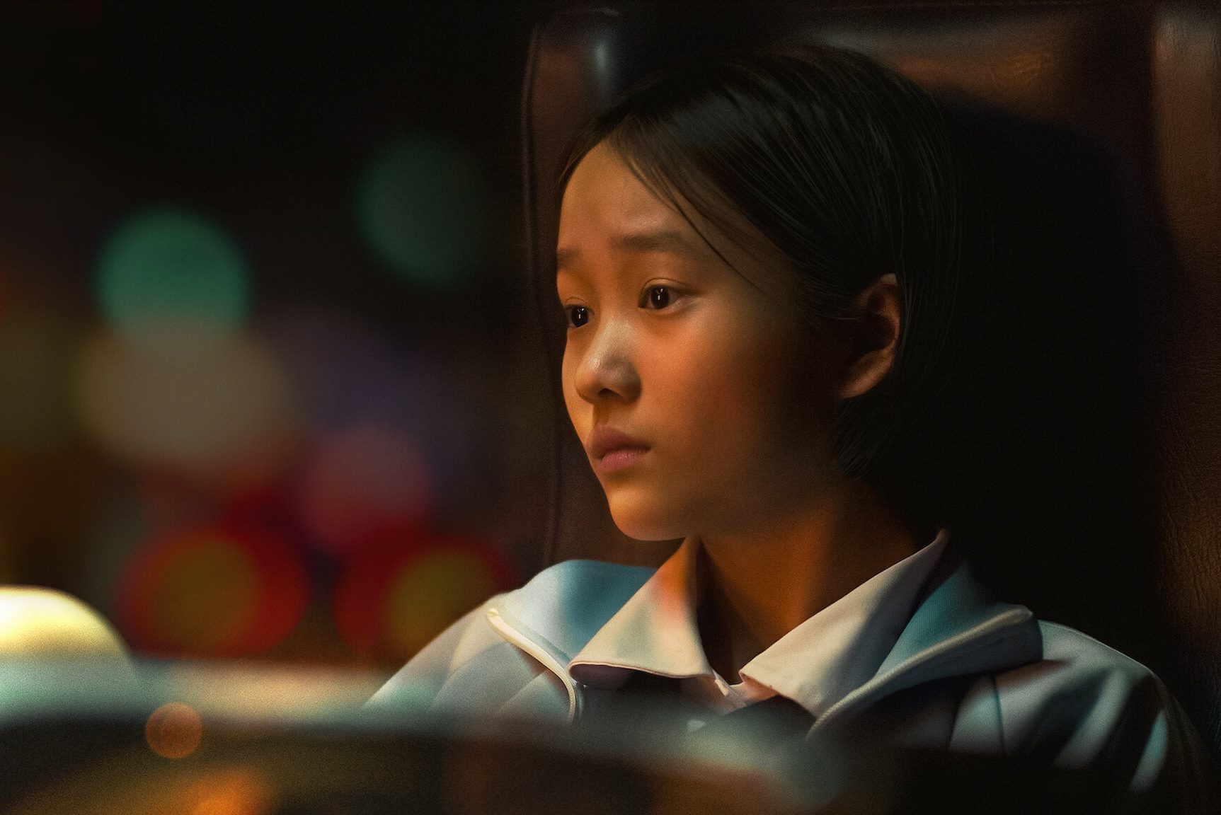 A girl sits pensively in a dimly lit room with colorful blurred lights in the background.