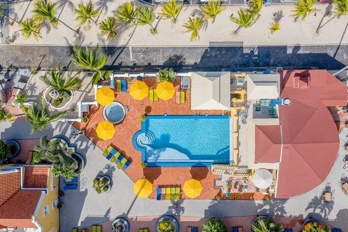 Swimming pool reflects sunlight, surrounded by yellow umbrellas and lounge chairs, amidst palm trees and a red-roofed building in a resort setting.
