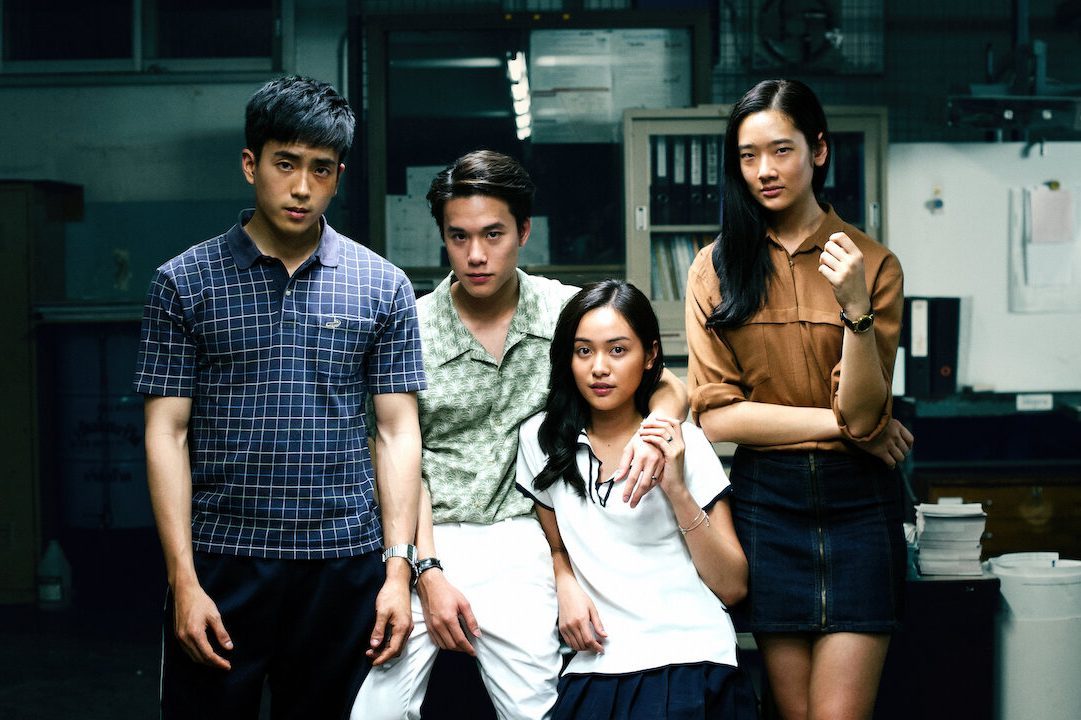 Four people stand confidently in an office-like setting, with serious expressions, surrounded by various office equipment and filing cabinets.