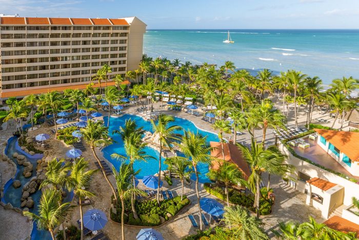 A hotel overlooks a large pool surrounded by palm trees and umbrellas near the ocean, with a sailboat visible on the horizon.