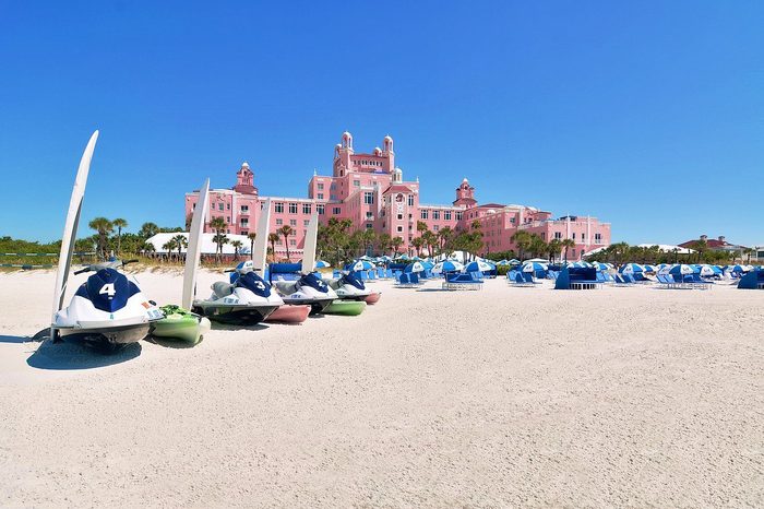 Jetskis rest on sandy beach; background features a large, pink building with palm trees and blue canopies under clear blue sky.