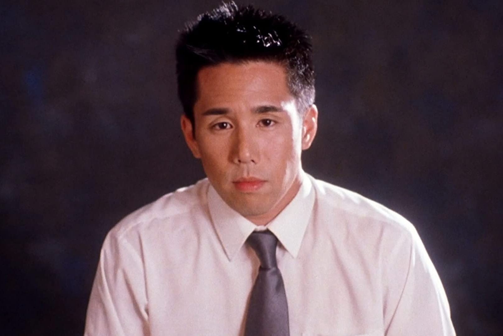 Person wearing a white shirt and gray tie, staring forward against a dark, blurred background.