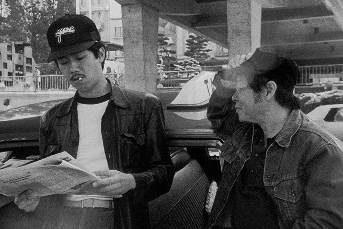 Two men stand by a car; one reads a newspaper with a cigarette, the other adjusts his cap. Urban setting with buildings in background.