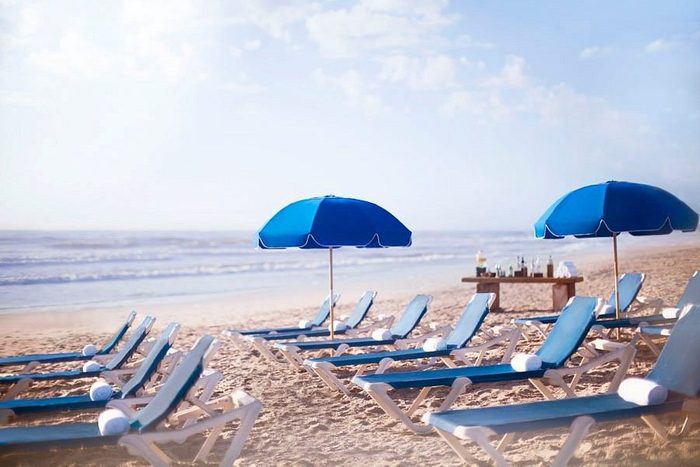 Blue lounge chairs and umbrellas line the sandy beach, facing the ocean, with a small table of drinks nearby.