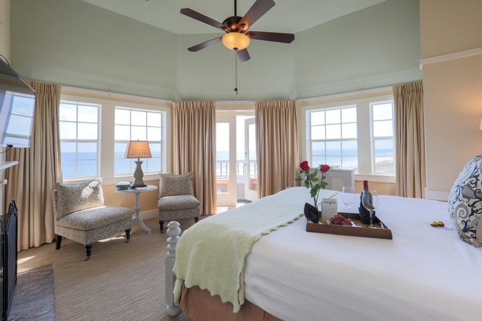 Bed holds tray with wine and glasses; roses placed near. Cozy room with chairs and table, large windows offer ocean view, ceiling fan above.