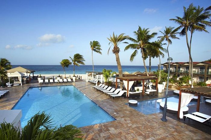 Pool surrounded by lounge chairs and cabanas, set against a background of palm trees and an ocean view under a blue sky.