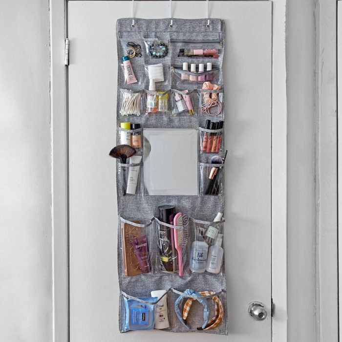 Organizer hangs on a door, holding various cosmetics and toiletries in clear pockets; a small mirror is in the center for convenience.