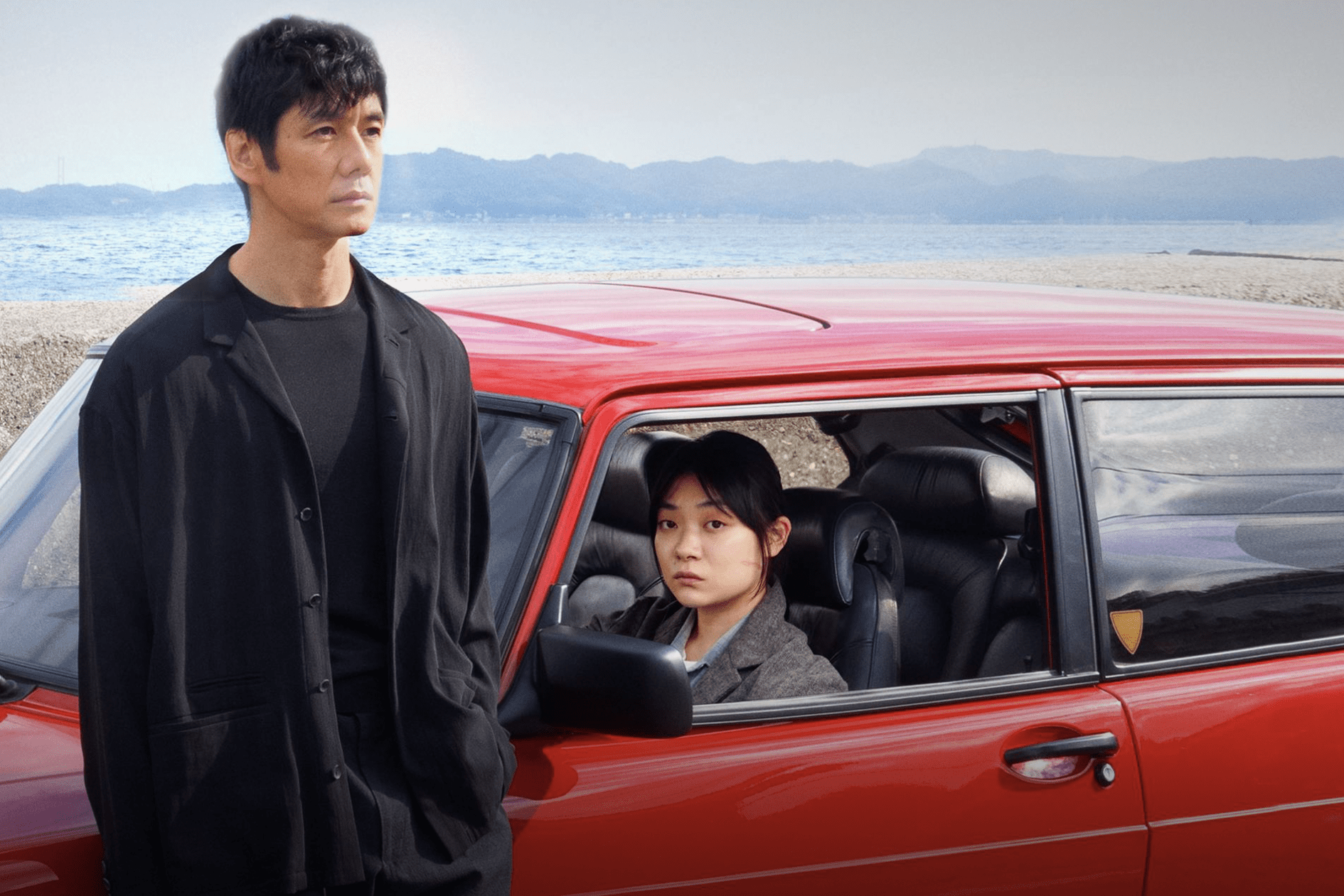 Person stands beside a parked red car, another person sits inside. The ocean and mountains form the background.