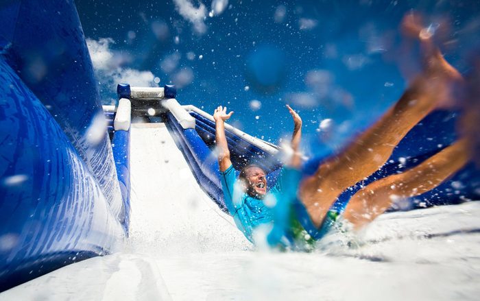 Person sliding down a blue water slide, arms raised, splashing water all around, under a clear blue sky.