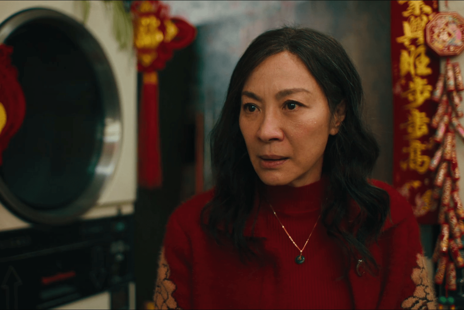 A woman stands thoughtfully, wearing a red sweater, surrounded by festive decorations and a washer in an indoor setting.