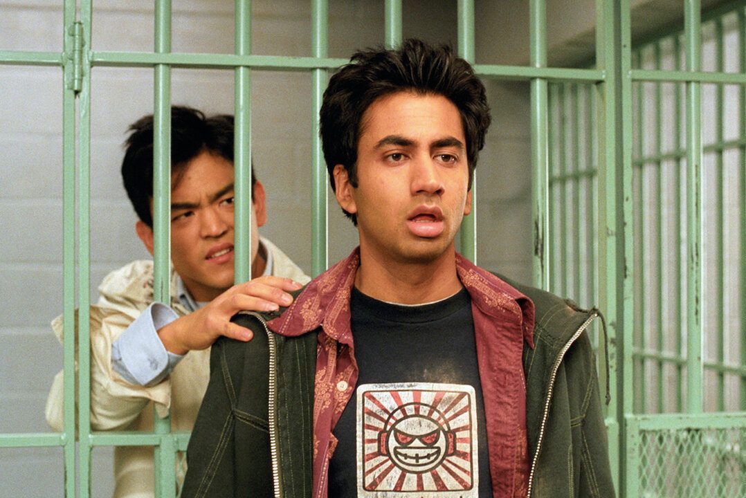 Two men stand in a jail cell, one looking surprised while the other grabs his shoulder. Bars and gray walls surround them.