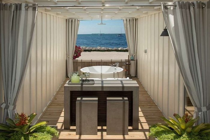 Bathtub sits in an open-air cabana, overlooking the ocean, with curtains, a wooden deck, and cocktail table, framed by tropical plants.