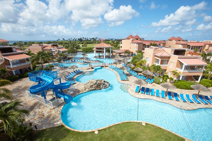 A blue waterslide descends into a large pool, surrounded by lounge chairs and thatched umbrellas, set amid resort buildings under a cloudy blue sky.
