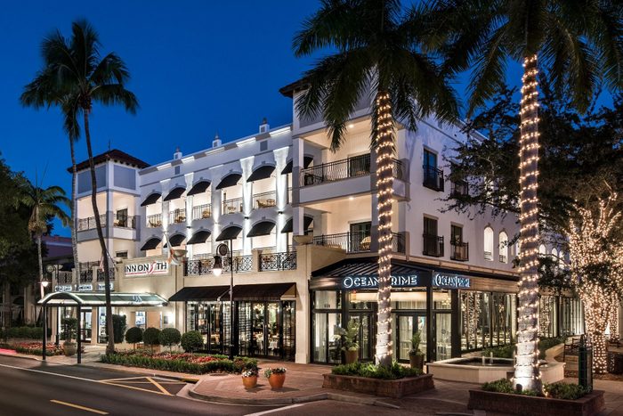 Hotel building illuminated at night, featuring balconies with awnings, palm trees wrapped in lights, located on a city street. Signs read 
