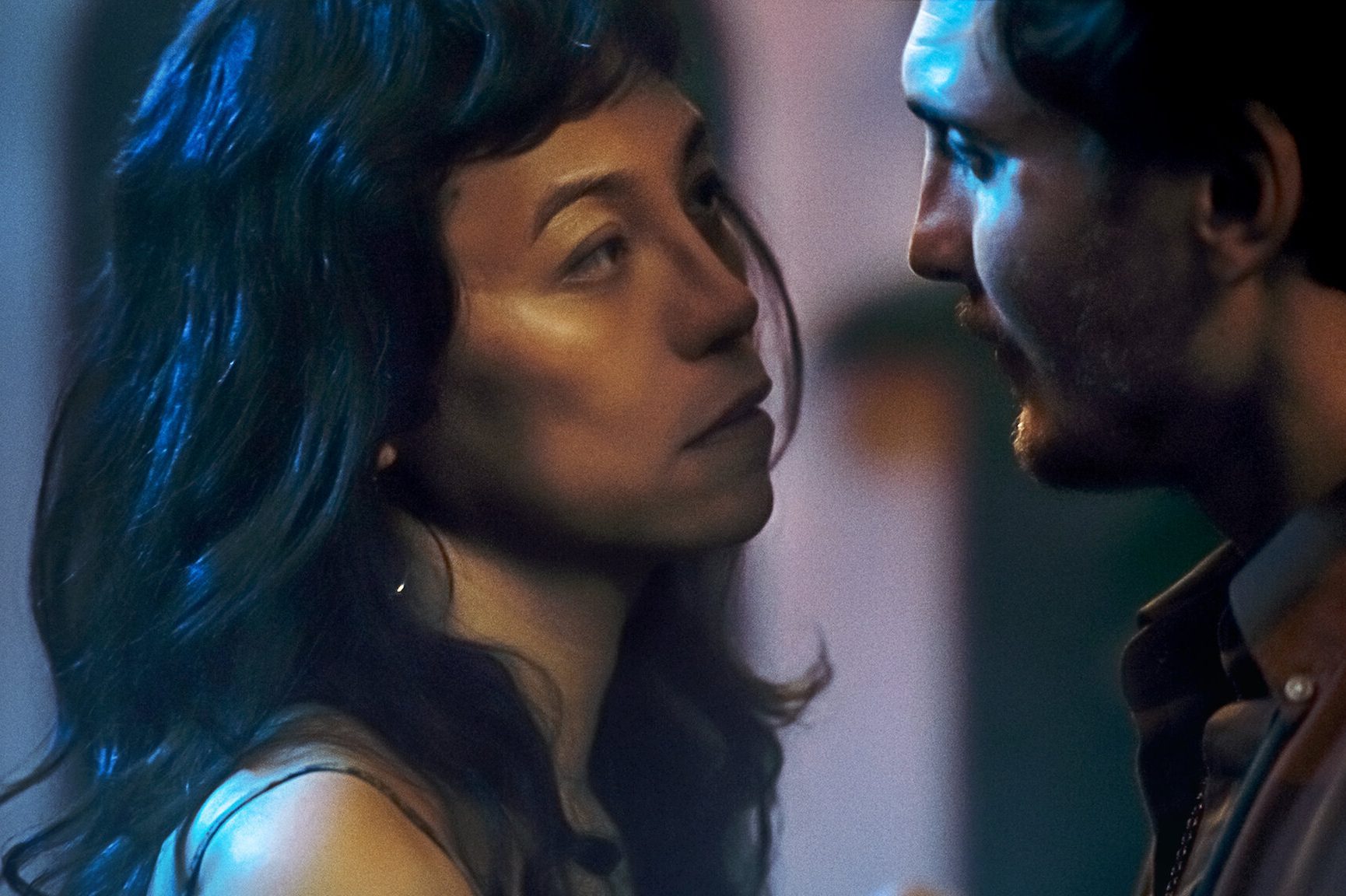 A woman and a man face each other closely, seemingly in a dim, intimate setting with soft lighting.