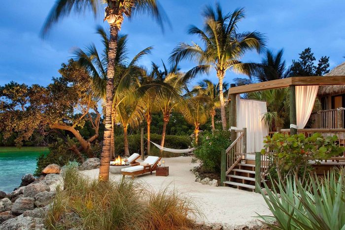 Lounge chairs face a fire pit on a sandy path lined with palm trees and a hammock, beside a cabana with curtains.